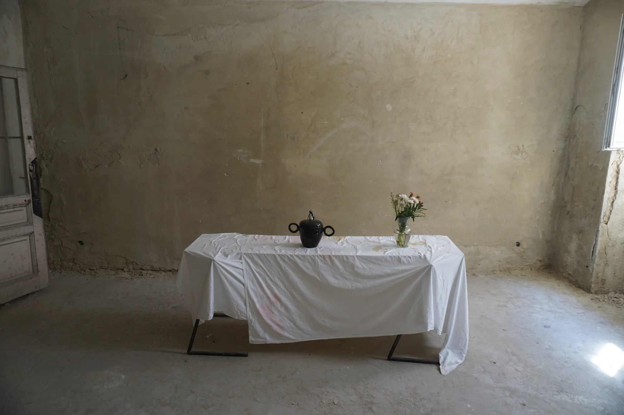 A table covered with a white sheet in a bare, vacant room, with a black ceramic pot and a vase of flowers on top