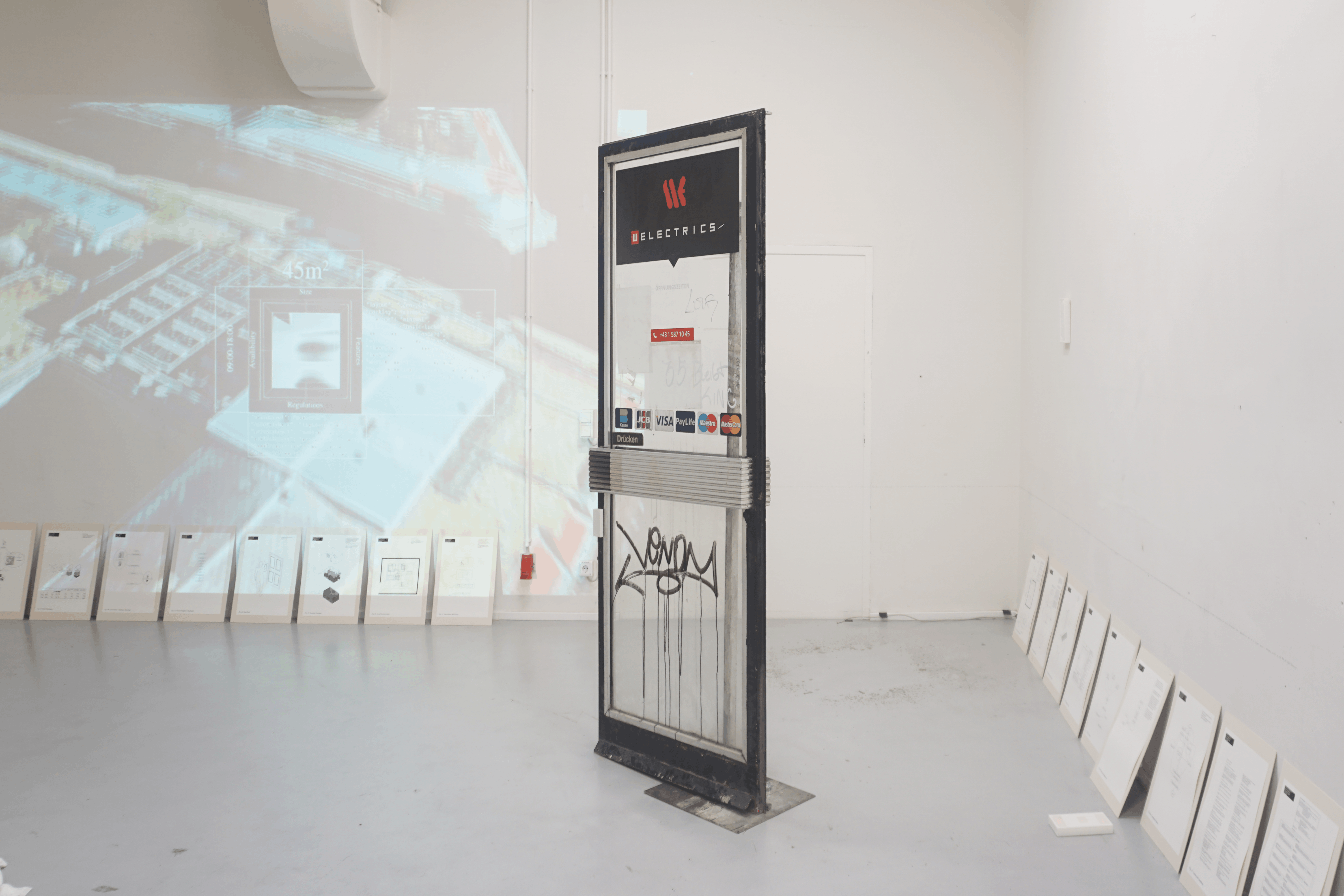 Installation view: a freestanding, graffitied shop door with card payment stickers in front of a projected aerial view, with framed sheets leaning along the walls