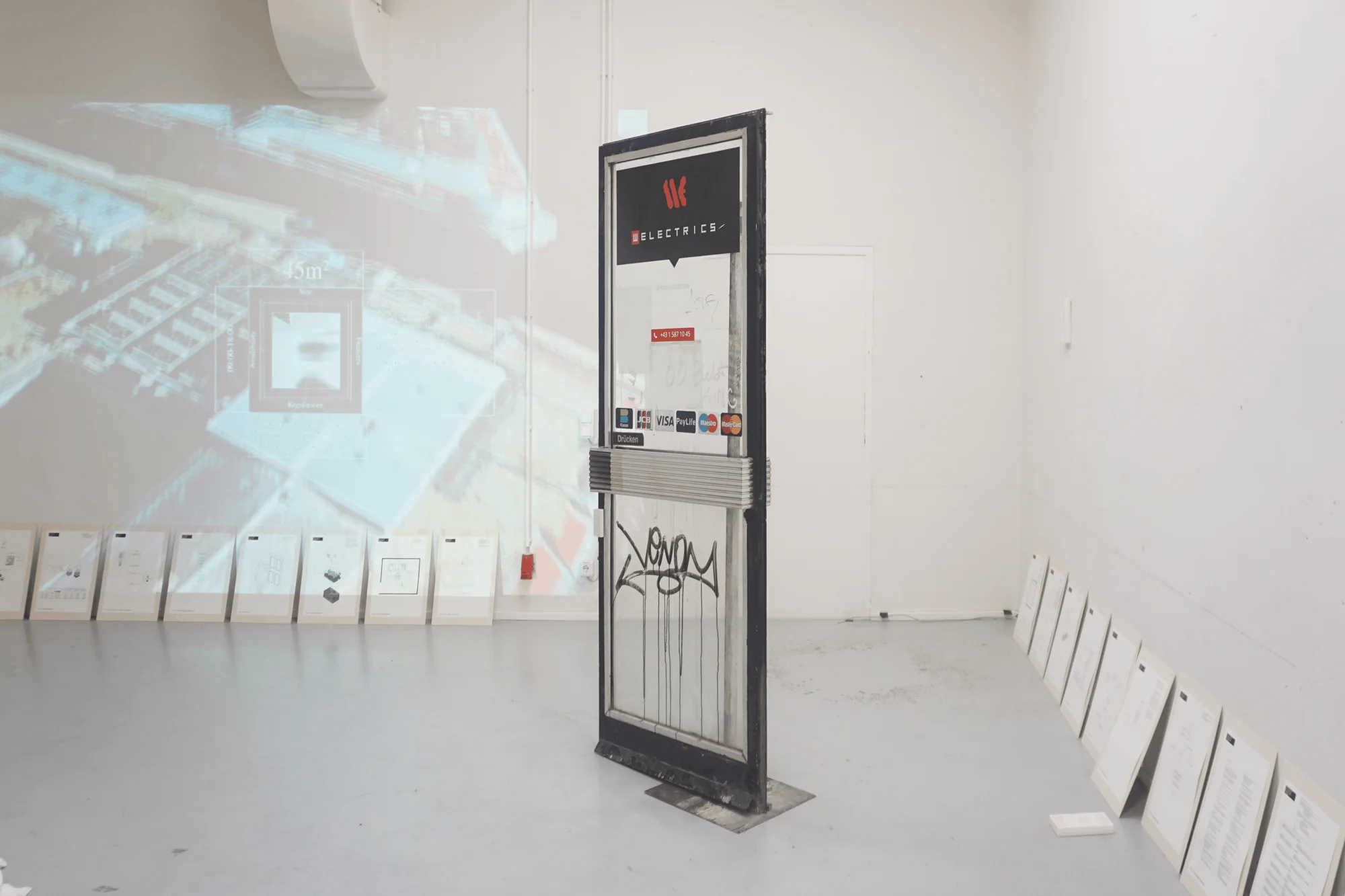 Installation view: a freestanding, graffitied shop door with card payment stickers in front of a projected aerial view, with framed sheets leaning along the walls