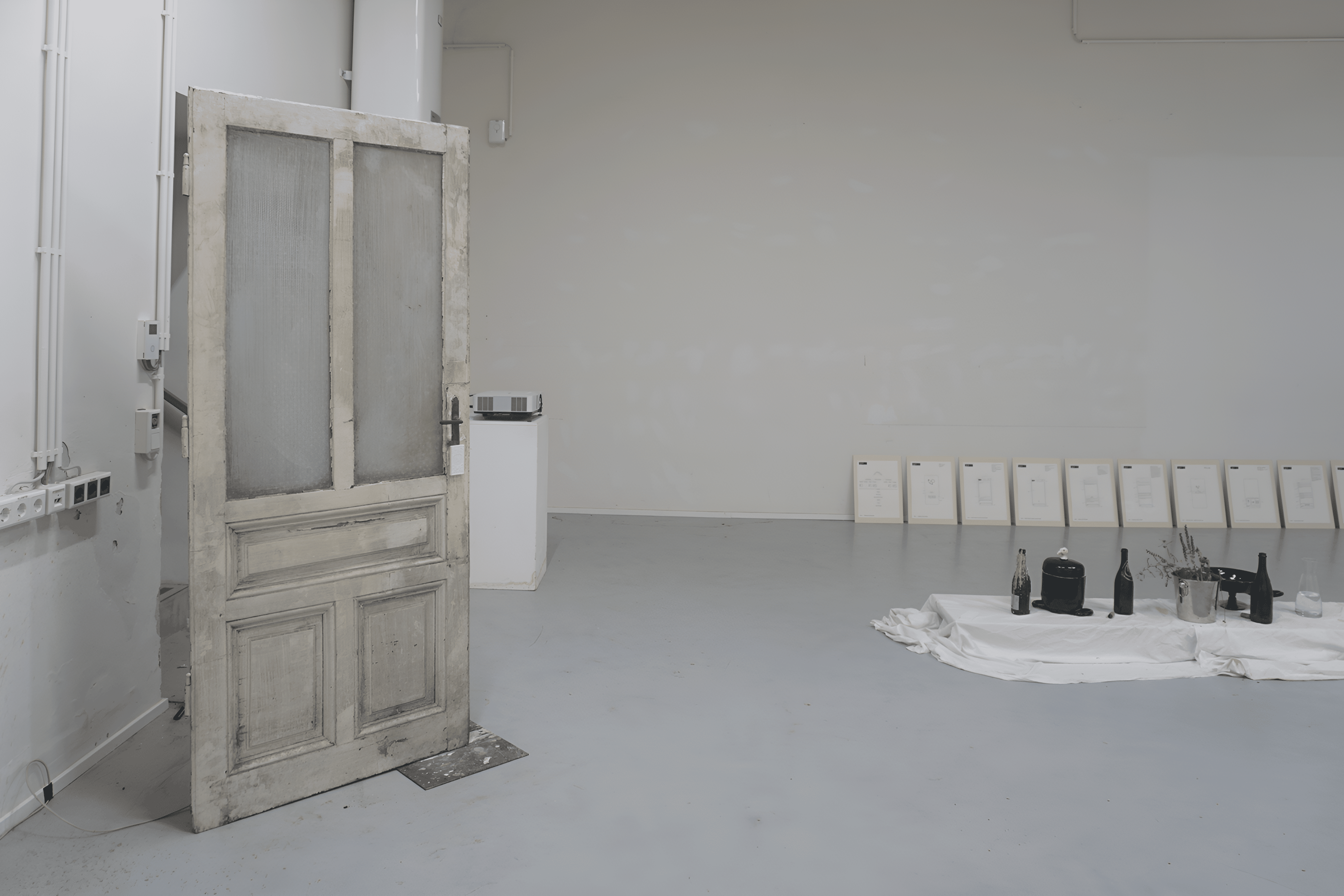 Installation view: a weathered wooden door standing open in a white room, a white cloth with black bottles and a plant on the floor and framed sheets leaning on the back wall
