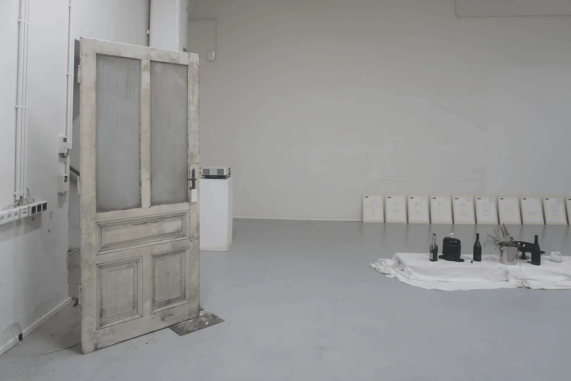 Installation view: a weathered wooden door standing open in a white room, a white cloth with black bottles and a plant on the floor and framed sheets leaning on the back wall