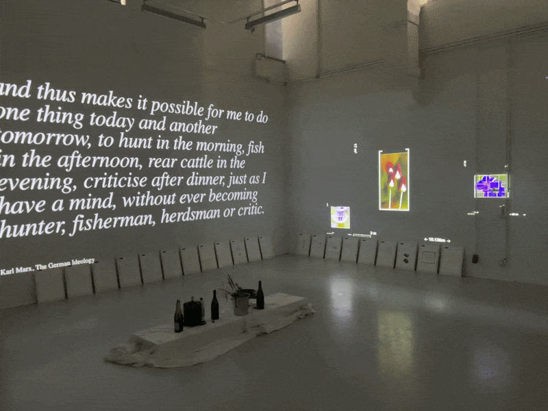 Installation view: a projected quote from Karl Marx's The German Ideology on one wall, then the quote 'The world consists of events, not things', framed sheets leaning along the walls and a white cloth with bottles and a plant on the floor