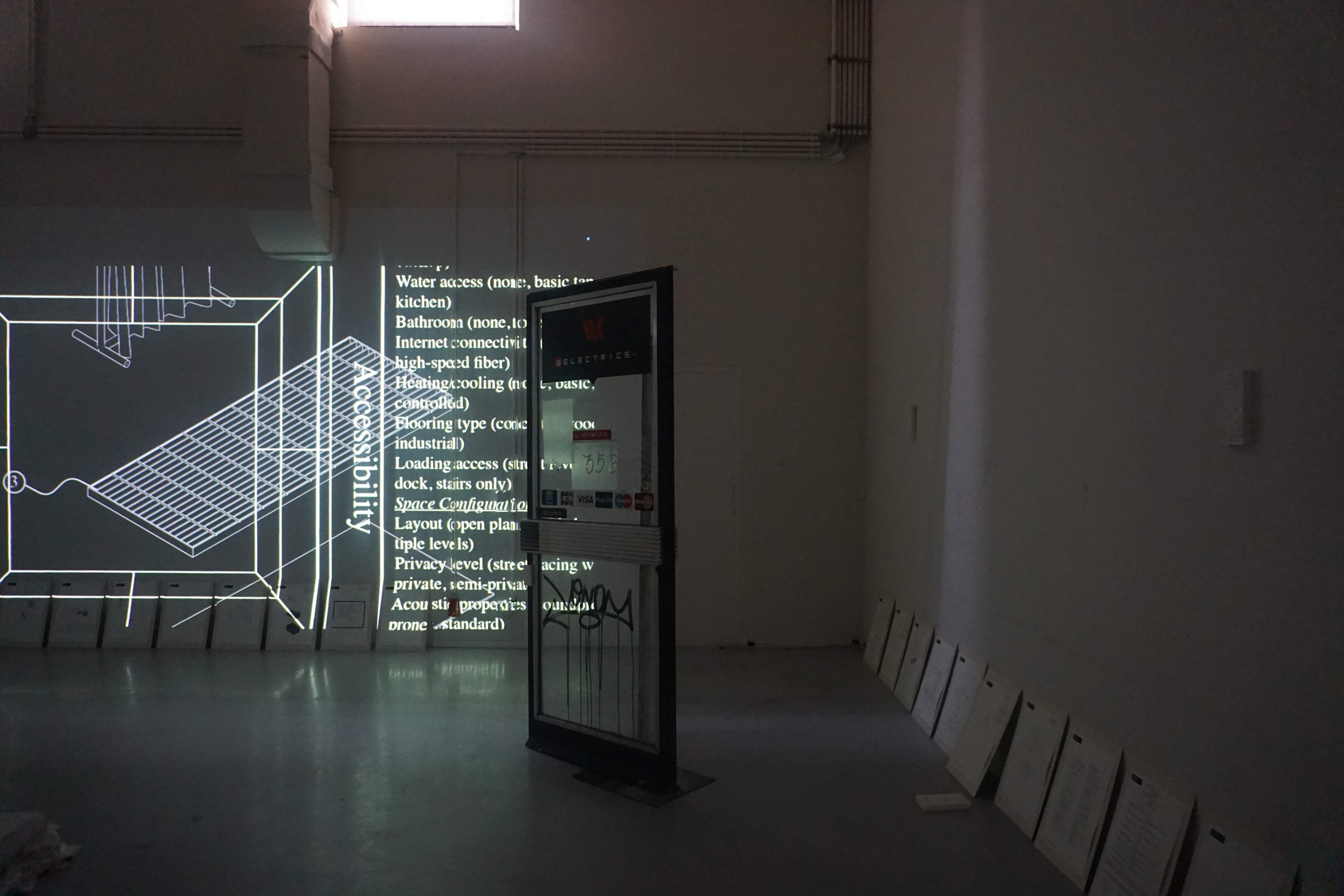 Dim installation view: a projected axonometric drawing and a list of space criteria such as accessibility, water and internet next to a freestanding shop door with graffiti